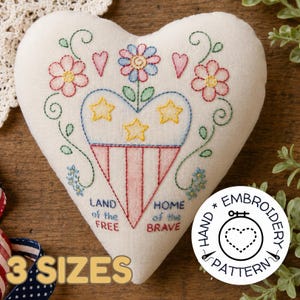 May include: Heart-shaped, off-white embroidered pillow with patriotic design. Features red, white, and blue stitching with floral and star motifs. Text reads "Land of the Free" and "Home of the Brave". Includes "3 Sizes" text.