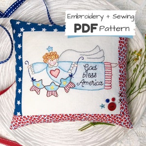 Op de afbeelding: Een wit kussen met een rood, wit en blauw geborduurd ontwerp van een engel die een banner vasthoudt met de tekst "God bless America". Het kussen heeft een blauwe en witte sterrenpatroonrand en een rode en witte bloemenpatroonrand.