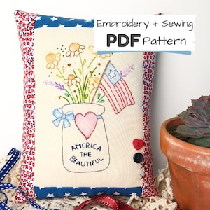 May include: Embroidered pillow with a red, white, and blue American flag, a mason jar filled with flowers, and the text "America the Beautiful".