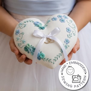 Puede incluir: Un cojín para anillos blanco en forma de corazón con bordados florales azules y turquesas, un lazo de satén blanco y dos anillos dorados. Un logotipo circular muestra el texto HAND EMBROIDERY SEWING PATTERN con iconos de corazón y máquina.