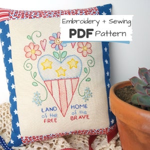 Könnte beinhalten: Ein weißes Kissen mit roter, weißer und blauer Stickerei eines Herzens mit Sternen und den Worten "Land of the Free" und "Home of the Brave". Das Kissen ist umgeben von rotem, weißem und blauem Stoff mit Sternen.