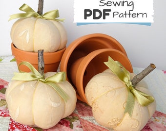 May include: Three cream-colored fabric pumpkins with green ribbon and twig stems, sitting in terracotta pots on a pink and green patchwork quilt. The image includes the text "Sewing PDF Pattern".