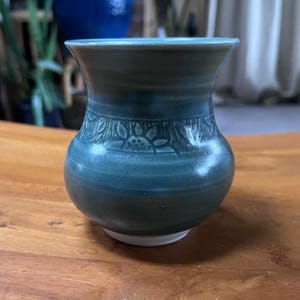 May include: A handcrafted teal ceramic vase with a wide opening and a rounded body. The vase features a decorative band of embossed floral patterns around the middle. The base is white. The vase is sitting on a wooden surface.