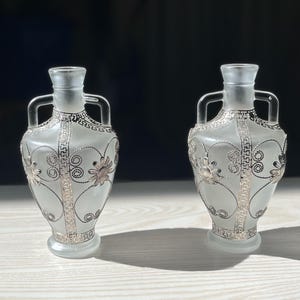 May include: Two frosted glass vases with silver-colored floral and geometric designs. Each vase has a narrow neck, two handles, and a decorative base. The vases are displayed on a light-colored surface against a dark background.