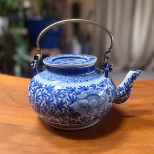 May include: A blue and white porcelain teapot with a floral design. The teapot has a rounded body, a curved spout, and a metal handle. The lid is decorated with a blue and white pattern. The teapot is sitting on a wooden surface.