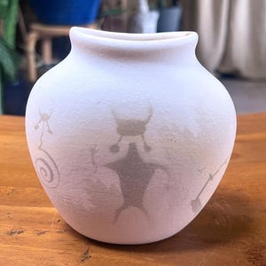 May include: A white ceramic vase with a rounded body and a slightly flared rim. The vase features faded gray illustrations of figures and symbols, including a central humanoid form with outstretched arms and a horned head. The vase sits on a wooden surface.