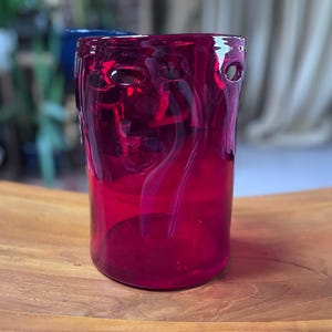 May include: A vibrant, ruby-red glass vase with a cylindrical shape. The vase has several circular openings near the top and a smooth, glossy finish. The vase is displayed on a wooden surface, showcasing its rich color and design.