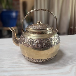 May include: A brass teapot with intricate embossed designs. The teapot features a curved spout, a hinged lid, and a handle. The body of the teapot has a band of detailed relief carvings. The teapot is sitting on a light-colored wooden surface.