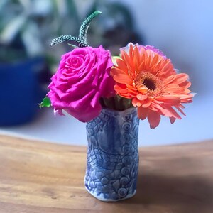 May include: A small, blue ceramic vase with a textured pattern holds a vibrant arrangement of flowers. The arrangement includes a bright pink rose, an orange gerbera daisy, and a decorative stem. The vase sits on a wooden surface.