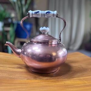 May include: A copper teapot with a white and blue ceramic handle and lid knob. The teapot has a curved spout and a rounded body. The handle is attached to the body with copper supports. The teapot is sitting on a wooden surface.