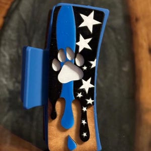 May include: A blue and black hair clip with a paw print and white stars. The clip has a blue stripe and dripping blue paint design. The base of the clip is a light brown color.