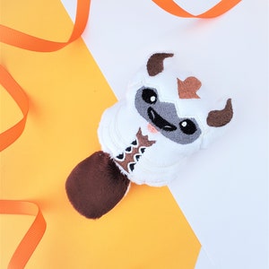 May include: White plush toy resembling a yak, with brown fur and a brown and white embroidered design on its chest. The toy is on a yellow and white background.