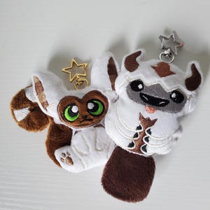 May include: Two plush keychains of animated characters. One is a white and brown flying bison with a smiling face. The other is a brown and white lemur with large green eyes. Both have star-shaped key rings attached.