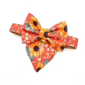 Può includere: Un papillon arancione vibrante con un motivo a girasoli con fiori gialli e arancioni e foglie verdi. Il papillon è fissato con una chiusura color oro e ha un cinturino arancione abbinato con un design floreale.