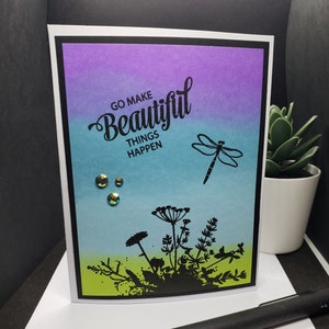 May include: Greeting card with a gradient background of purple, teal, and green. The card features the text "GO MAKE BEAUTIFUL THINGS HAPPEN" and a dragonfly silhouette. Embellished with iridescent gems and a floral silhouette.