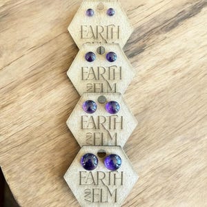 Peut inclure: Quatre paires de boucles d'oreilles en améthyste sur des cartes en bois hexagonales avec le texte "Earth and Elm" imprimé sur chaque carte.