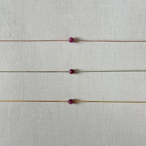 May include: Three delicate necklaces with a single, small, faceted, dark red gemstone pendant on each. The necklaces are shown against a white background. The necklaces are made with different coloured chains: rose gold, silver, and gold.
