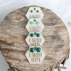 May include: Three pairs of green and blue gemstone stud earrings on hexagonal cards with the text "Earth and Elm" printed on them.