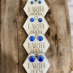 Puede incluir: Cuatro pares de pendientes de botón de lapislázuli azul mostrados en tarjetas hexagonales con el texto "Earth and Elm" impreso en cada tarjeta.