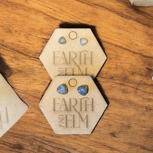 May include: A pair of iridescent blue gemstone earrings on a hexagonal card with the text "Earth and Elm".