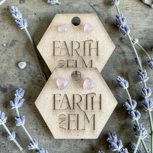Könnte beinhalten: Zwei Paar rosafarbene Edelstein-Ohrstecker, präsentiert auf sechseckigen Holzanhängern. Die Anhänger sind mit den Worten "EARTH AND ELM" graviert. Lavendelzweige und kleine Steine umgeben die Ohrringe.