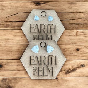 May include: Two light blue triangle earrings sit on a hexagonal wooden display. The display is engraved with the words "EARTH AND ELM". The background is a wooden surface.