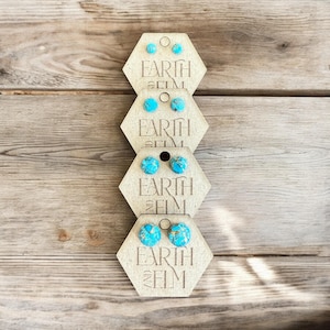 May include: Four pairs of turquoise stud earrings presented on hexagonal wooden cards. The cards are engraved with the words "EARTH AND ELM". The earrings feature a speckled pattern and are set on a wooden surface.