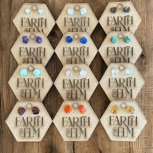 Peut inclure: Neuf paires de boucles d'oreilles présentées sur des cartes en bois hexagonales. Chaque carte porte le texte "EARTH AND ELM" et présente une paire de boucles d'oreilles différente avec des pierres précieuses de différentes couleurs, notamment bleu, vert, violet, orange et transparent.
