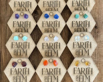 May include: Nine pairs of earrings displayed on hexagonal wooden cards. Each card features the text "EARTH AND ELM" and showcases a different pair of earrings with various gemstone colors, including blue, green, purple, orange, and clear.