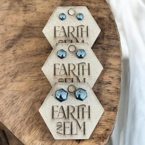 May include: Three pairs of blue stud earrings displayed on hexagonal wooden cards. Each card is engraved with the words "EARTH AND ELM". The earrings have a faceted, geometric design and are set on a wooden surface.
