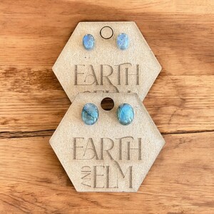 Peut inclure: Deux paires de boucles d'oreilles à clous avec des pierres ovales bleu-vert sont présentées sur des cartes hexagonales de couleur beige. Les cartes portent le texte "EARTH AND ELM". Les boucles d'oreilles sont présentées sur une surface en bois.