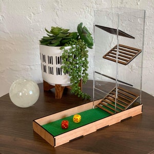 Könnte beinhalten: Ein Würfeltower aus Holz mit einem klaren Acrylturm und einem grünen Filztablett. Zwei Würfel sind im Tablett sichtbar, einer gelb und einer rot.