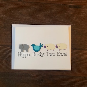 以下が含まれることがあります： 水色の縁取りのある白いグリーティングカードには、灰色のカバ、青い鳥、クリーム色の羊が2匹描かれています。「Hippo, Birdy, Two Ewe!」というテキストが黒いフォントで書かれています。カードは濃い茶色の木製の表面にあります。