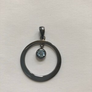 May include: A silver pendant with a black finish featuring a round circle with a light blue gemstone hanging from the center.
