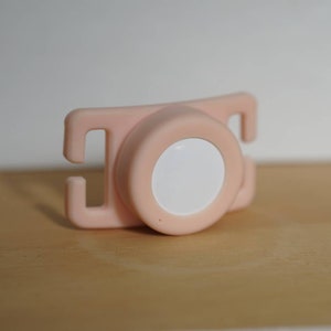 May include: A pink plastic safety strap with a white button in the center. The strap has two loops on either side.