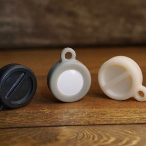 May include: Three round, plastic keychains with different colors and designs. The keychain on the left is black, the middle keychain is gray with a white circle in the center, and the keychain on the right is white.