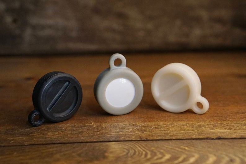 May include: Three round, plastic keychains with different colors and designs. The keychain on the left is black, the middle keychain is gray with a white circle in the center, and the keychain on the right is white.
