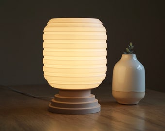 May include: A white ceramic table lamp with a pleated shade and a beige base. The lamp is turned on and illuminating the room. A small white vase with a green plant is on the table next to the lamp.