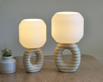 May include: Two white ceramic table lamps with a textured, ring-shaped base and a white frosted glass shade. The lamps are turned on and illuminated.