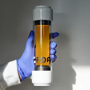 Könnte beinhalten: Ein durchsichtiger Glaszylinder mit weißem Ober- und Unterteil, der eine gelbe Flüssigkeit enthält. Der Zylinder hat ein schwarzes Etikett mit dem Text "RDA".