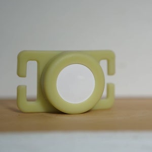 May include: A light green plastic buckle with a white circle in the center. The buckle is designed to be used with a strap or belt.