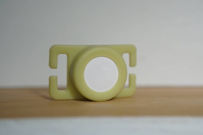 May include: A light green plastic buckle with a white circle in the center. The buckle is designed to be used with a strap or belt.