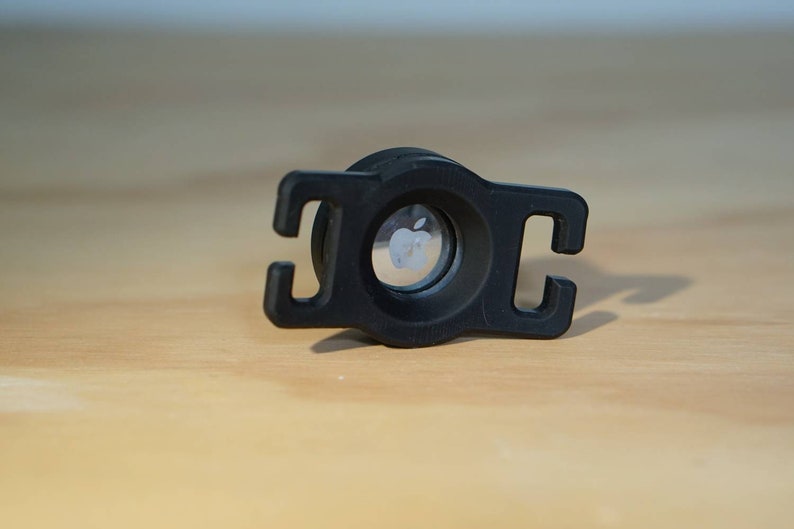 May include: Black plastic adapter with a circular opening featuring the Apple logo. The adapter has two loops on either side.