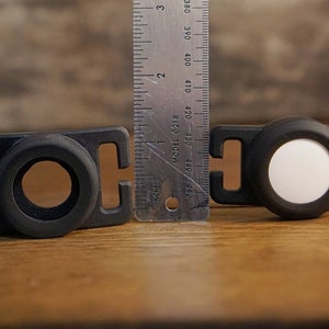 May include: Two black plastic clips with a white reflective disc on one and a black ring on the other. The clips are about 1 inch wide and are next to a ruler.