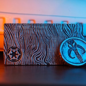 May include: A rectangular, textured metal object with a wood grain pattern and an Imperial symbol, next to a round coin with a Mandalorian symbol. The objects are silver and gray, with a blue and orange gradient background.
