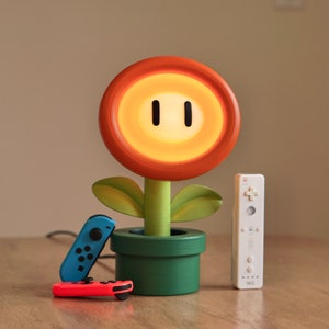 May include: A wooden lamp shaped like a Piranha Plant from the Super Mario video game series. The lamp is orange, green, and yellow and has a light-up face. The lamp is sitting on a wooden table with a blue and red Nintendo Switch controller and a white Wii remote.