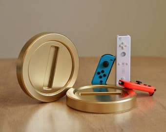 May include: A golden coin prop with a large "1" cutout, next to a smaller coin and three game controllers. The controllers are blue, white, and orange. The items are arranged on a wooden surface.