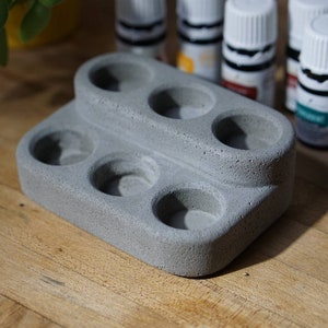 May include: A gray concrete holder with six round indentations for essential oil bottles. The holder has two tiers and is made of a rough, textured material.