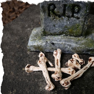 May include: A collection of white bone props, resembling human bones, are scattered on a gray, textured surface. A weathered tombstone with the inscription "RIP" is visible in the background. Tombstones sold separately.