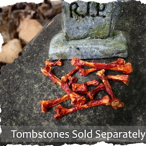 May include: A pile of fake, red-stained bones scattered on a grey, textured surface in front of a small, grey tombstone with the inscription "RIP". Tombstones sold separately.